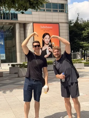 Dois amigos fazendo coração com os braços numa praça de Seul
